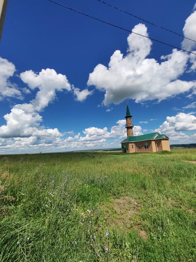 This image depicts a mosque with a minaret, situated in a grassy field under a bright blue sky dotted with white, fluffy clouds. The mosque is constructed of light-colored wood and has a green roof. The minaret is tall and slender, also topped with a green spire, and has several small windows. The surrounding landscape is a vast expanse of green grass, with some wildflowers visible. In the distance, the land appears to roll gently, suggesting a rural or semi-rural setting. A power line runs across the upper portion of the image. The scene is bathed in natural light, indicating it is daytime, likely mid-morning or afternoon, given the bright sky. The weather appears to be clear and pleasant. The overall impression is one of tranquility and open space, with the religious structure standing as a focal point in the natural environment. There is no visible text in the image.