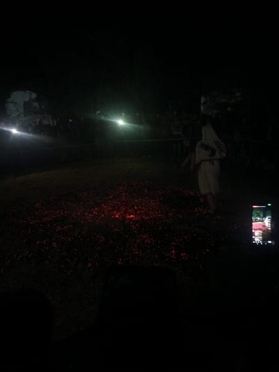 In the city of Dennitsa, Bulgaria, a dark, outdoor scene unfolds at night, likely during a cultural event or ritual. The ground is covered in what appears to be a bed of glowing embers or hot coals, emitting a reddish-orange glow that illuminates the immediate surroundings. In the center of this fiery path, a person dressed in a long, white garment is walking. This individual is moving from right to left within the frame, towards the left side of the image, with their back mostly to the viewer. The fabric of their attire is flowing, suggesting movement.

A crowd of spectators is gathered around the periphery of the fiery area, mostly visible as silhouettes against the darkness. Their presence indicates an audience observing the central action. Several bright lights, possibly from torches or artificial lighting, are strategically placed, casting harsh beams and creating lens flares that further obscure details in the background and highlight the central performance. On the right side of the image, a smartphone is being held up, likely recording the event, with its screen displaying a bright, colorful image, possibly a live feed of the scene itself. The overall environment is characterized by darkness punctuated by the intense glow of the coals and artificial lights, creating a dramatic and somewhat mystical atmosphere.