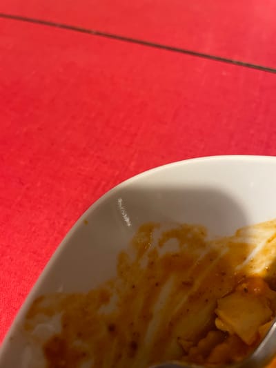 A close-up depicts a partially eaten meal on a white plate, with remnants of food and sauce smeared across its surface. The plate is placed on a red surface. A utensil is partially visible, suggesting recent use. The image focuses on the immediate aftermath of dining.