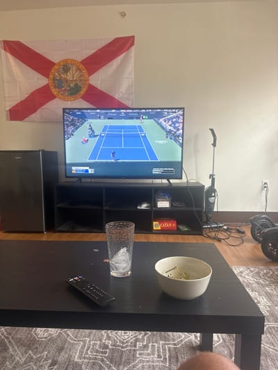 An indoor scene in Miromar Lakes, United States, depicts a living room. A large flat-screen television, positioned on a black entertainment stand, displays a tennis match on a blue court with two players and spectators. Above the television, a Florida state flag is affixed to the light-colored wall. To the left of the TV stand, a black Midea mini-fridge is present. To the right of the stand, a black stick vacuum cleaner stands upright, and two black, two-wheeled hoverboards are partially visible. A tangle of black electrical wires is on the light wooden floor in this area. In the foreground, a black rectangular coffee table rests on a patterned rug. On the coffee table, a clear glass tumbler with ice, a black remote control, and several scattered coins are visible. To the right of the glass, a light-colored bowl contains several small, greenish-brown objects. The lower portion of a human leg is partially visible in the bottom right corner of the image.