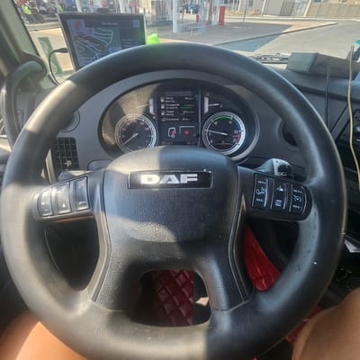 The image shows the interior of a DAF truck cab, viewed from the driver's perspective. The steering wheel, bearing the DAF logo, is prominent in the foreground. The dashboard displays various gauges, including a speedometer and tachometer, and a digital screen showing fuel consumption data. On the left side of the steering wheel are media control buttons. On the right side are cruise control buttons. A GPS navigation device is mounted on the windshield. Outside the truck, a gas station with multiple pumps and vehicles is visible. The scene appears to be at a gas station in La Palme, France, as indicated by the "La Palme" text on the GPS screen. The lighting suggests daytime. No individuals are clearly visible within the cab.