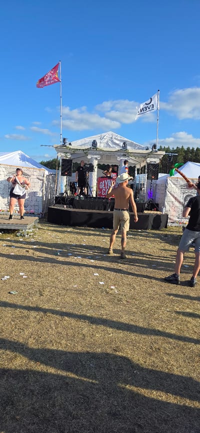 The image depicts an outdoor festival scene in Neustadt-Glewe, Germany. A stage, resembling a Greek temple facade, is set up with speakers and lighting. Several individuals are on and around the stage, including a DJ. To the left, a woman is performing on a smaller elevated platform. In the foreground, a man wearing a cowboy hat and shorts is facing away from the camera. The ground is covered with dry grass and scattered debris, suggesting a public gathering or event. Two flags are visible in the sky above the stage.