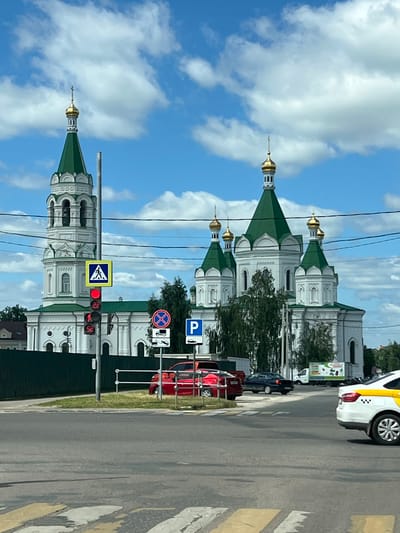 A low-angle, eye-level, full shot captures a large, white church with green, domed roofs against a bright blue sky filled with fluffy white clouds. The church features multiple domes, the tallest of which is on a bell tower to the left, topped with a gold cross. In the foreground, a crosswalk is visible on an asphalt road, with traffic lights and road signs. A pedestrian crossing sign is illuminated red, with a countdown timer showing "22" and "52". To the right of the traffic lights, a red car is parked, and further down the road, a blue sedan is in motion. A white and yellow taxi is also visible on the right side of the image. The scene appears to be in a city, with a green fence bordering the church property. The location is identified as Yegoryevsk, Russia.