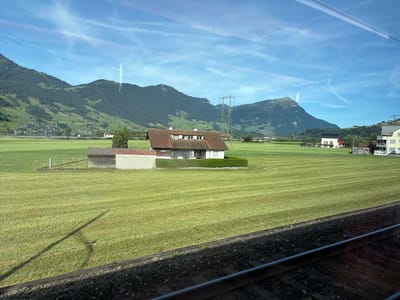 The image depicts a rural landscape with a house and outbuildings situated in a vast, meticulously mowed green field. In the background, rolling hills covered in dark green trees ascend towards a bright blue sky with wispy clouds. A large electricity pylon stands prominently on one of the hills. In the far distance, a small cluster of buildings can be seen, hinting at a village or town. The foreground shows a section of railroad tracks, suggesting the vantage point is from a moving train. The overall scene conveys a serene, pastoral setting in Switzerland. The location is Steinen, Switzerland.