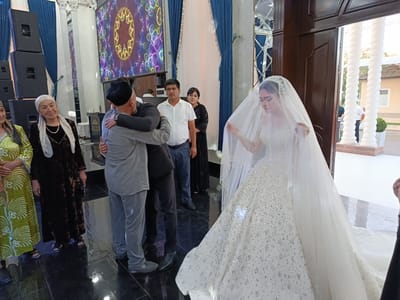 In a brightly lit indoor venue in Shahrisabz City, Uzbekistan, a wedding celebration is underway. The scene captures a poignant moment as a man in a grey shirt and pants, wearing a dark kufi, embraces another man in a dark suit, also wearing a kufi. Their hug appears warm and affectionate, suggesting a familial or close bond.

To the right of the embraced men, a bride in a dazzling white wedding gown, adorned with sparkling embellishments, stands partially obscured by her veil. Her gaze is directed downwards, hinting at a sense of solemnity or shyness. The elaborate gown trails on the polished dark floor.

In the background, several guests are visible. On the left, three women are dressed in traditional attire. One woman wears a vibrant green outfit with intricate white patterns and a headscarf. Next to her, another woman is in a dark, patterned dress with a white headscarf. A third woman, partially visible, wears a similar dark, patterned outfit.

Further back, a man in a white polo shirt and blue jeans stands attentively, while a woman in a black dress is positioned behind him. The venue is decorated with ornate pillars, draped blue curtains, and a large screen displaying a colorful, abstract pattern of hearts. Large black speakers are stacked on the left side of the room. The overall atmosphere suggests a joyous but also respectful occasion. The presence of traditional clothing and the ornate decor point to a culturally significant event, likely a wedding ceremony or reception. The lighting indicates it is daytime or a well-lit indoor evening event. No visible text is present in the image.