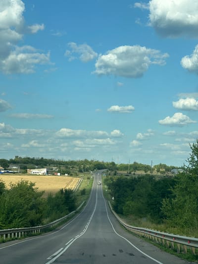 The image depicts a rural landscape with a paved road curving through rolling hills. The road is lined with a metal guardrail on the right side, and the surrounding vegetation consists of green trees and bushes, with a field of dry, golden crops to the left. In the distance, there are a few small buildings, possibly farm structures. The sky is a bright blue with scattered white cumulus clouds. Several vehicles are visible on the road, including cars and a truck. The overall scene suggests a sunny day in a countryside setting.