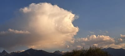 This image captures a broad, panoramic view of a landscape under a partly cloudy sky. The prominent feature is a large, voluminous cumulus cloud, tinged with the warm hues of late afternoon or early morning sunlight, dominating the left and central portions of the sky. To the right, smaller, more dispersed cumulus clouds drift across a clear blue sky. The horizon is marked by a range of dark, silhouetted mountains. In the foreground, the tips of several dark green bushes or trees are visible, adding a touch of vegetation to the scene. The overall impression is one of natural beauty and the vastness of the sky. The location is identified as Bash-Kaiyndy, Kyrgyzstan.