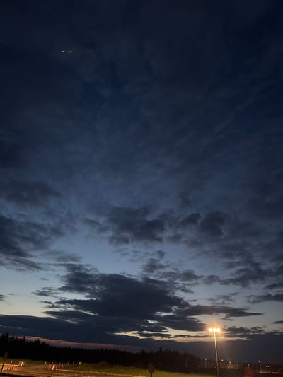 This is a photograph of a dark, cloudy sky at dusk. The sky is a deep blue with patches of lighter blue and gray clouds. In the distance, there is a faint glow of light on the horizon, likely from the setting sun. The bottom of the image shows a silhouetted line of trees, and below that, a dimly lit road or parking lot with some streetlights. There are no people visible in the image. The location is identified as Stony Plain, Canada.