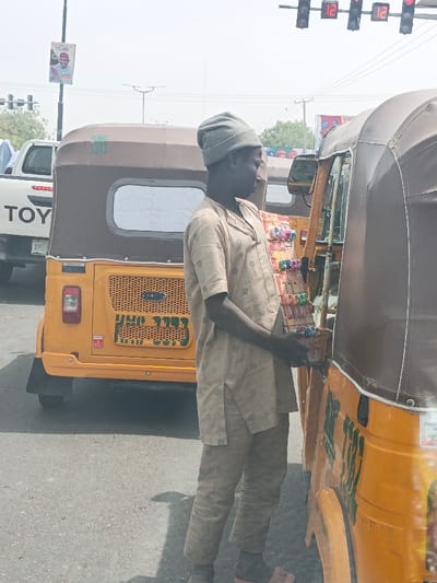 The image captures a daytime street scene in Hotoro, Nigeria. In the foreground, a male individual with dark skin, wearing a grey knit cap and a light-colored, short-sleeved patterned tunic with matching trousers, stands in profile facing right. He holds a rectangular display rack containing numerous small, packaged items, positioned alongside a yellow and brown motorized tricycle (rickshaw).
In the midground, a second yellow and brown motorized tricycle is visible, featuring a rear panel with the alphanumeric identifier "KMG 3373" in green lettering, and a "FORD" logo. Behind this tricycle, a white Toyota pickup truck is partially visible, displaying "TOYOTA" on its tailgate. The vehicles are situated on an asphalt road.
Overhead, a set of three vertical traffic lights displays red signals, with two featuring a digital countdown timer showing "12". A tall street pole in the upper left quadrant of the image bears a political poster depicting a man in a cap, accompanied by text. The sky appears hazy or overcast.