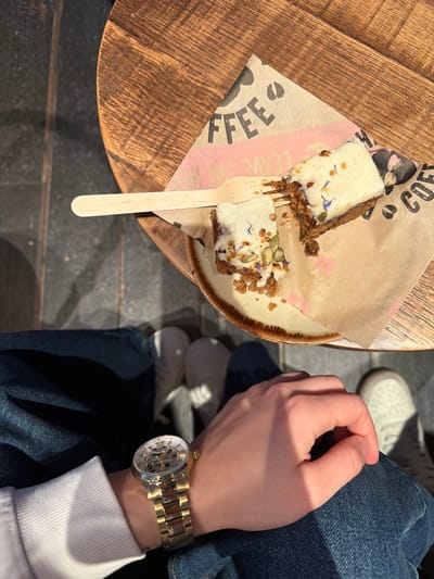 The image provides an overhead view of a person seated at a round, light-brown wooden table in an establishment located in Bretton, United Kingdom. On the table, a ceramic plate with a brown rim holds two pieces of a baked good, likely a cake, topped with white frosting, pumpkin seeds, and small blue botanical elements. The cake is situated on light-brown parchment paper marked with the word "COFFEE" and a coffee bean graphic. One piece of cake shows evidence of consumption, with a section removed and crumbs scattered on the plate and paper. A light-colored wooden fork rests on the paper, partially touching the cake.
The lower half of a person is visible beneath the table. The individual is wearing blue denim jeans and a white long-sleeved garment on their left arm. A gold-toned automatic skeleton watch with Roman numerals and a metallic link bracelet is worn on the left wrist, with the left hand resting on the left knee. On the dark-colored floor directly beneath the table, two feet are visible, clad in light-colored athletic shoes or sneakers. Strong directional lighting from the upper right casts distinct shadows from the table and the person's legs onto the floor.