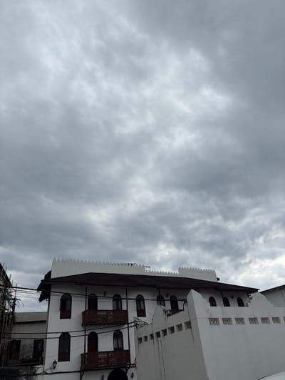 In Kelele Square, Tanzania, a three-story white building with arched windows and wooden balconies stands under a cloudy sky. The building has a dark tiled roof and a decorative crenellated parapet. To the left of the building, scaffolding is visible, suggesting ongoing construction or renovation. A lower white wall with small, rectangular openings runs in front of the main building. Electrical wires are strung across the scene. The sky is filled with heavy, gray clouds.