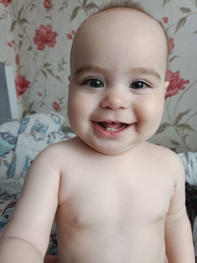 A close-up, eye-level shot shows a smiling baby's face and upper chest. The baby has dark eyes, light skin, and sparse light brown hair on their head. Their mouth is open, revealing two front teeth. The baby is facing forward and appears to be looking directly at the camera. The background is a floral wallpaper with pink and red flowers on a light background. To the left of the baby is a white and blue blanket with owl illustrations. The lighting is soft and natural. The image was taken in Noviy, Russia.