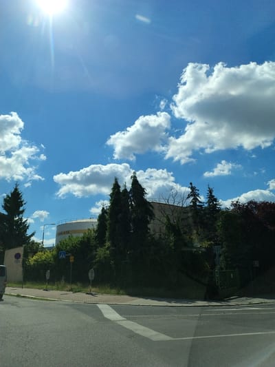 The image shows a street scene in Strakonice, Czechia, on a sunny day. The foreground consists of a road with a pedestrian crossing. To the right of the road, there is a sidewalk with several traffic signs and a green fence. In the background, there are trees, a large white cylindrical structure with yellow lettering that reads "BOŘÍ MLÝNY", and a bright blue sky with scattered white clouds. The sun is prominently visible in the upper left corner of the frame, casting a lens flare. A portion of a white van is visible on the far left of the image.