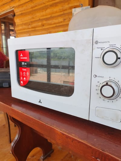 The image shows a white Accessstyle microwave oven placed on a wooden table. The microwave is positioned at an angle, with its front facing the viewer and slightly to the right. The oven door is closed, and the glass panel reveals a grid-like interior. On the left side of the oven door, a red sticker displays "accesstyle," "20 л," and icons representing different cooking functions, including "Соло" (Solo). The right side of the microwave features two control dials. The upper dial is labeled "Мощность" (Power) and has settings for "Средняя" (Medium) and "Низкая" (Low), along with a defrosting icon. The lower dial is labeled "Таймер" (Timer) and has markings for time in minutes, from 0 to 35. The microwave appears somewhat dusty.
The table is made of dark, polished wood with a reddish-brown hue, and it has an ornate, curved support structure. The background consists of a wooden structure, possibly a wall or ceiling, constructed from horizontal logs. The overall lighting suggests an outdoor or semi-outdoor environment, likely during the daytime.
The context of Voskresensky District, Russia, is not visually discernible from the image itself, as the scene lacks specific landmarks or identifying features related to that location. The image depicts a static scene focusing on the microwave and its immediate surroundings.