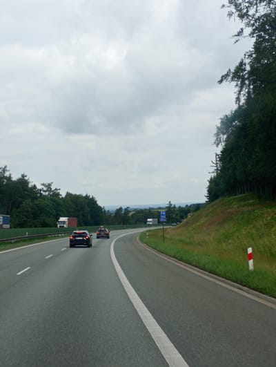 The image captures a view from a moving vehicle on a highway in Góra Świętej Anny, Poland, under an overcast sky. The highway has at least two lanes visible, with white lane markings. In the foreground, the road surface is dark asphalt. On the right side of the road, a grassy embankment rises, topped by a dense line of trees. Red and white striped markers are visible along the edge of the embankment. In the middle ground, two dark-colored passenger cars are driving ahead on the highway. To the left, a white truck and a red trailer are parked, with a blue and white sign on a blue barrier in the far left. A blue sign with white text, indicating "PA 262 500m", is also visible to the right of the highway. The background shows a distant landscape of trees and potentially some structures, fading into the hazy sky.