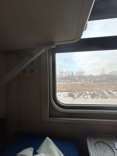 The image depicts the interior of a train car, observing an external winter landscape near Usolye-Sibirskoye, Russia, during daytime.
Inside the train, a light-colored, hinged overhead bunk or storage shelf is partially lowered, supported by a beige articulating arm. Below it, on a beige wall panel, two square QR codes with Russian text are affixed, positioned above a small, round silver button. A dark grey, rounded window frame encloses the view outside, its glass displaying visible smudges. In the lower portion of the image, a dark blue fabric surface, likely a seat or lower bunk, holds light-colored, knitted garments. To the bottom right, a silver-textured surface features a circular indentation, indicative of a cup holder or integrated table.
Outside the window, a winter scene unfolds under an overcast sky. Multiple parallel train tracks are visible in the immediate foreground, with patches of snow covering the ground between them. Beyond the tracks, a strip of dry, brown vegetation is interspersed with irregular snowdrifts. A long, low concrete wall spans the mid-ground. Above and behind this wall, leafless deciduous trees and some evergreen foliage are visible, with distant buildings and overhead utility lines appearing on the horizon. No people are visible within the frame.