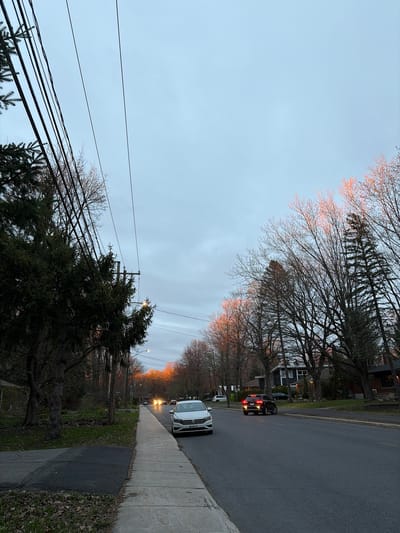 This media file captures a quiet suburban street scene at either dawn or dusk, likely in Saint-Bruno-de-Montarville, Canada. The sky is a soft, pale blue-grey, with a warm, orange glow illuminating the tops of bare trees in the distance, characteristic of the golden hour.
The asphalt road stretches from the foreground into the background, lined on the right by a concrete sidewalk. A white SUV, possibly a Volkswagen, is parked on the right side of the street in the mid-ground. Further down the road, a dark-colored car shows illuminated red taillights, and multiple bright headlights from other vehicles are visible in the far distance, indicating low light conditions.
Both sides of the street are bordered by numerous trees, predominantly deciduous with bare branches, suggesting late autumn, winter, or early spring. Some evergreen trees are also visible, particularly in the foreground and near a utility pole on the left. Behind the trees, glimpses of residential houses can be seen.
A prominent utility pole with several crisscrossing power lines and communication cables stands on the left side of the image. At least two streetlights are visible, one in the mid-ground on the right and another further down the road, appearing to glow faintly. The ground alongside the sidewalk features patches of green grass interspersed with fallen brown leaves.
No specific activity or people are visible; the scene depicts a static, tranquil moment. The Volkswagen logo is visible on the front of the white SUV, but no other discernable text can be read, such as on license plates. The overall ambiance is peaceful and still, bathed in the soft, transitional light of daybreak or twilight.