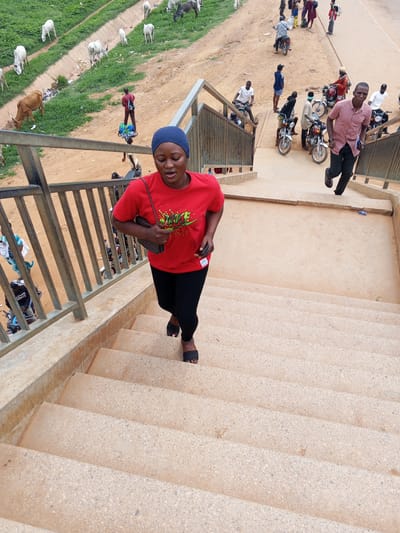 A woman wearing a blue headscarf and a red t-shirt with a graphic print walks up a concrete staircase. She carries a dark patterned handbag. She is wearing black leggings and black sandals. Behind her, a man in a pink shirt and dark pants walks down the stairs, holding a book or notebook. In the background, several motorcycles are parked, and people are gathered around them. The scene appears to be outdoors, with a dusty, reddish-brown ground. To the left of the stairs, there is a grassy slope with several white and brown cattle grazing. The sky is overcast, suggesting a cloudy day. The overall atmosphere appears busy, with a mix of pedestrian and motorcycle activity. The location is identified as Sauka, Nigeria. There is no visible text in the image.