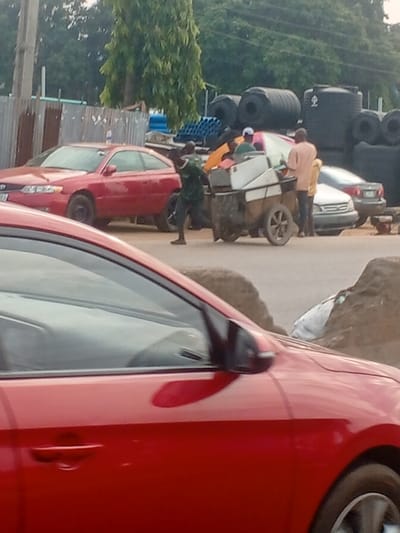 The image depicts a roadside scene in Abule Egba, Nigeria, under diffused daylight. In the immediate foreground, the right side of a red sedan is partially visible, indicating the viewpoint from another vehicle.

In the midground, a red two-door coupe is parked or stationary on an unpaved shoulder adjacent to a corrugated metal fence. To its right, three individuals are clustered around a large, two-wheeled, rudimentary cart. The cart features a white upper section and a dirt-stained lower body, carrying various items including a white appliance (possibly a refrigerator unit) and colorful textiles (orange, pink, green). A male in a dark green shirt and dark trousers is positioned to the left of the cart, appearing to push or pull it. Another male, wearing a light brown shirt, stands to the right rear of the cart. A third male, partially obscured, is also near the cart wearing a green shirt and possibly a light-colored cap.

Further down the unpaved road, a silver sedan is parked or stopped. The background on the left features a utility pole behind the metal fence and a tall, slender tree with green foliage. On the right, a commercial area is visible, characterized by large stacks of black, cylindrical plastic water tanks, bundles of blue pipes, and black tires, suggesting a building materials or plumbing supply establishment. Piles of dirt or debris are visible on the ground in the mid-right.