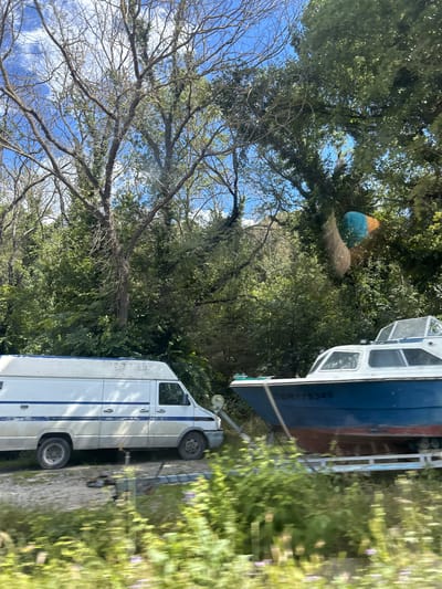 The image depicts an outdoor scene in Zvezditsa, Bulgaria, on a bright, sunny day. In the foreground, blurred green foliage with some purple flowers is visible, suggesting motion blur, possibly from a vehicle. To the left, a white panel van with blue trim is parked on a gravel surface. To the right, a blue and red boat is situated on a trailer, also on the gravel. Behind the vehicles and boat, a dense, lush green forest occupies the background. The sky above is a clear blue with some white clouds. The overall impression is of a semi-rural or industrial area adjacent to a wooded environment.