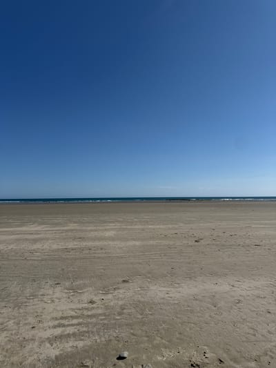 An outdoor, vertical, wide shot captures a tranquil beach scene under a clear blue sky. The foreground is dominated by a vast expanse of damp, sandy beach, showing subtle textures and the marks of previous footsteps or vehicle tires. A single grey pebble rests in the bottom right corner of the frame. In the mid-ground, the gentle waves of the ocean meet the shore, creating a thin line of white foam. A few scattered individuals are visible in the distance along the water's edge, appearing as small, dark silhouettes against the lighter sand and sea. The horizon is clearly defined by the meeting of the deep blue sea and the bright blue sky, with no visible landmass on the other side of the water. The overall impression is one of open space and natural serenity.