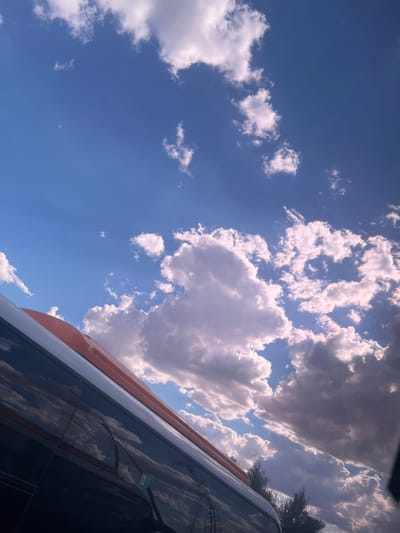 The image captures a low-angle view of a bright blue sky filled with fluffy white and grey clouds. Sunlight filters through, creating bright patches in the sky and casting shadows on some of the clouds. The scene is viewed from the perspective of being inside or next to a vehicle, with the reflective surface of a tinted window occupying the lower right portion of the frame. The window reflects the sky and clouds, as well as a glimpse of trees and power lines in the distance. A portion of the vehicle's exterior is visible along the top edge of the window, featuring an orange and white stripe. A green sticker with some faint text is visible on the window. The overall impression is of a sunny day, possibly late morning or early afternoon, with a sense of movement as if traveling. The setting appears to be outdoors, consistent with a location in Satbayev, Kazakhstan.