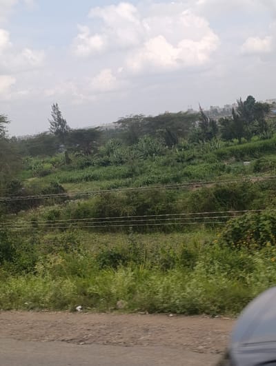 The image depicts a verdant landscape in Matungulu west ward, Kenya, under an overcast sky with scattered white clouds. The foreground shows a dirt and gravel road surface, with a dark grey vehicle component partially visible in the bottom right. The midground features dense green vegetation, including shrubs and taller trees, with several horizontal utility lines running across. An upward sloping terrain, heavily covered in lush green growth and interspersed with trees of various species, dominates the scene. In the distant background, at a higher elevation, several structures and buildings are visible, indicating urban or semi-urban development. No individuals are present in the scene.