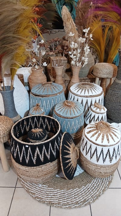 This is a close-up, eye-level, medium shot of a retail display of woven baskets and decorative dried floral arrangements. The items are arranged on a patterned woven rug placed on a tiled floor. The background is cluttered with more woven baskets, vases, and dried grasses. No people are visible. The style of the decor suggests an artisanal or bohemian aesthetic, possibly found in a boutique or market stall. The mention of Bergerac, France, in the prompt is not supported by any visual information within the image itself.