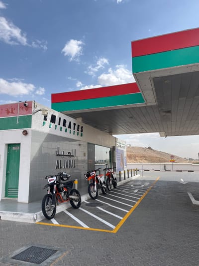 An Emirates Petroleum gas station with a red and green canopy stands under a cloudy sky. Four orange and black dirt bikes are parked in front of the station, next to white stripes on the dark pavement marked with yellow lines. A mountain range is visible in the background.