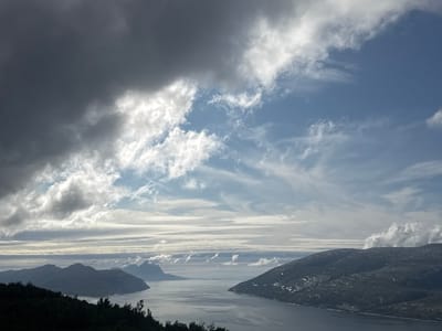 The image is a panoramic view of a fjord landscape in Flostrand, Norway. In the foreground, a dark, silhouetted treeline covers the bottom of the frame. Beyond this, a body of calm, grey water, likely a fjord, stretches into the distance. Several mountainous islands, shrouded in varying degrees of shadow and light, rise from the water. The sky is dramatic, with a mix of dark, heavy clouds on the left and lighter, wispy clouds against a blue sky on the right. The overall scene suggests a cool, possibly windy day with changing weather conditions.