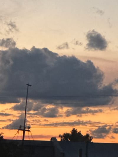 An outdoor, ground-level, medium shot captures a dramatic sky with scattered dark clouds silhouetted against a bright orange and light yellow sunset. The horizon is obscured by the dark silhouettes of rooftops and trees. A tall utility pole with wires and crossbars stands prominently in the mid-ground, breaking the expanse of the sky. The image appears to be a photograph taken in the city of Opochka, Russia, at dusk.