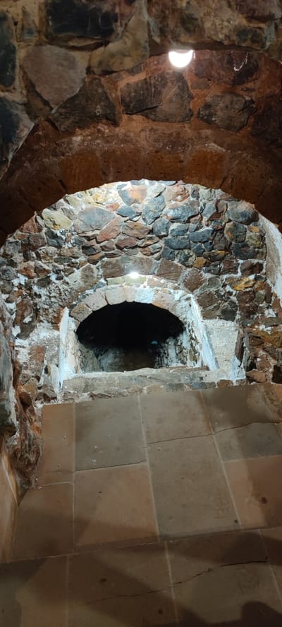 This image depicts the interior of an ancient stone structure, likely a historical oven or fireplace, located in Taperakan, Armenia. The scene is characterized by rough-hewn stone walls and arched openings. In the foreground, large, square tiles form a floor or step leading up to a smaller, arched opening that appears to be the entrance to the oven. The walls surrounding this opening are constructed from irregular stones of varying colors and sizes, cemented together with a reddish-brown mortar. Some white residue, possibly salt or mineral deposits, is visible on the stone surfaces. Above the oven opening, another larger stone arch frames the entire structure. Natural light sources, possibly bulbs, are visible illuminating the interior of the stone structure from above, casting a warm glow. The overall impression is one of age and rugged construction.
