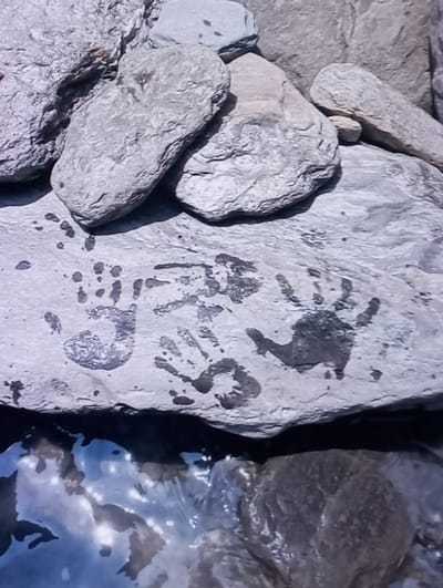 The image displays a close-up, elevated view of a rock formation adjacent to a body of water in Aktash, Uzbekistan. The upper portion of the frame shows an arrangement of stacked, irregular-shaped, light gray rocks with rough, weathered surfaces, varying in size. Below these, a larger, flatter, horizontal gray rock serves as a primary surface. This prominent rock exhibits multiple dark, wet-appearing handprints, consistent with human hands. At least four distinct handprints are discernible, showing variations in clarity and completeness, suggesting they were made by hands pressed onto the surface. Below this handprint-adorned rock, a body of water occupies the lower section of the frame. The water is dark blue-gray, reflecting bright light sources from above, creating glistening patterns. Submerged rocks with dark, rounded forms are visible within the water, partially obscured by the surface reflections. No people are visible in the frame, but their recent presence is indicated by the handprints.