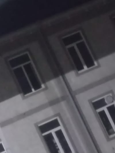 The image shows a blurry, low-light view of the facade of a building. The building appears to be two stories high, with multiple windows visible on each level. The windows are rectangular and have white frames. The exterior of the building is a light gray color. The image is tilted, suggesting it was taken from an angle. There are no people or any other discernible objects in the frame. The scene is likely an exterior view of a building at night or in very low light conditions. The specific location in Yorkatay, Uzbekistan cannot be determined from the visual information provided in the image.