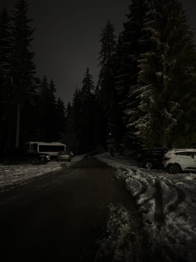 The image depicts a nighttime scene in what appears to be a wooded area in Пампорово, Bulgaria. A narrow road, partially covered in snow, cuts through the scene. On the right side of the road, there is a pile of snow. On the left side of the road, there are tall evergreen trees, also covered in snow. Two cars are parked on the side of the road. The lighting is dim, creating a dark and somewhat obscure view.