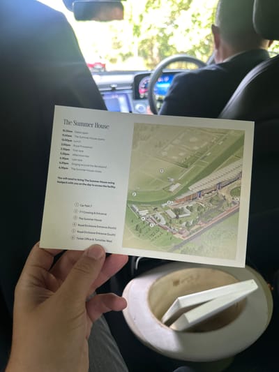 The image shows a person's hand holding a card with information about "The Summer House." The card has a schedule of events, including "Gates open" at 10:30 am and "The Summer House closes" at 6:30 pm. There is also a map of the grounds, with numbered locations such as "Car Park 7," "The Summer House," and "Royal Enclosure Entrance (North)."
The background suggests the person is inside a vehicle, possibly a car, with a driver in the foreground wearing a suit and tie. The environment appears to be outdoors, with trees visible through the car windows, indicating a daytime setting. The overall impression is that the person is preparing to attend an event at "The Summer House," possibly a racecourse, given the mention of races and enclosures on the card. The location of the Borough of Runnymede in the United Kingdom is relevant to the context of such events, as it is known for its historic sites and royal associations.