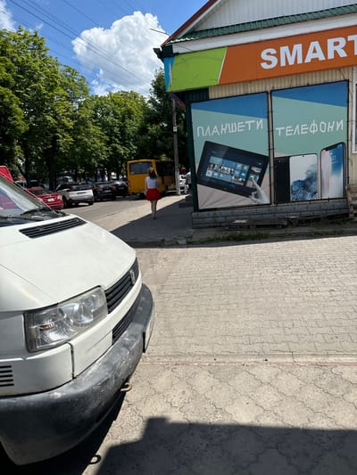The image depicts a street scene in Sarny, Ukraine. The setting is an outdoor commercial area with a building on the left displaying a large orange sign with white lettering that reads "SMART". Below this sign are two large banners advertising mobile devices. The top banner shows two smartphones and has Cyrillic text that translates to "TELEPHONES". The bottom banner features a tablet displaying a Windows interface and has Cyrillic text that translates to "TABLETS".
In the foreground, the front of a white van is visible, with a dark gray bumper. The van is parked on a paved surface. A woman in a red skirt and white top is walking away from the camera on the sidewalk. Behind her, a yellow van is parked, and further down the street, several other vehicles are lined up. Lush green trees border the street, and a bright blue sky with fluffy white clouds is visible above. The lighting suggests it is daytime, likely mid-morning or afternoon, with clear weather. The overall scene conveys a casual, everyday atmosphere of a commercial street.