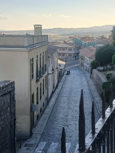 The image captures an elevated view of a cobblestone street in Ávila, Spain, winding downwards between historic stone and plaster buildings. The scene is bathed in the soft light of either early morning or late afternoon, indicated by the long shadows and the warm glow on the distant hills.
On the left side of the street, a prominent pale yellow building features multiple stories with regularly spaced windows and small balconies with dark metal railings. A stone wall, topped with what appears to be fragmented terracotta tiles, forms the immediate foreground on the left. On the opposite side, a narrower building with a red-tiled roof is visible, followed by other structures of varying heights and architectural styles. One building undergoing renovation is partially covered with a blue net.
The street itself is paved with tightly packed cobblestones, showing signs of wear. Several streetlights with a traditional design are mounted on the buildings. Two dark-colored trash receptacles are placed along the right side of the street. In the distance, the landscape is characterized by rolling hills and agricultural fields under a hazy sky. A metal fence with sharp, pointed finials runs along the bottom right edge of the frame, suggesting a viewpoint from a higher elevation or a balcony. No people are visible in the scene. The overall impression is one of a quiet, historical urban setting.