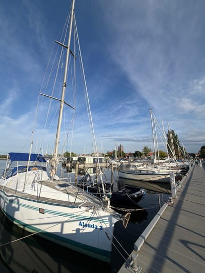 The image displays a marina scene with several sailboats docked along a wooden pier. The sky is blue with scattered white clouds, indicating fair weather. The sun casts shadows on the pier and the boats, suggesting it is daytime, likely late morning or afternoon. In the foreground, a white sailboat named "Alauda 2" is prominently featured. It has a green stripe along its hull and a blue cover over part of its deck. Multiple other sailboats with tall masts and rigging are lined up behind it, some with people on board or visible in the background. Further back, buildings and trees can be seen, providing context for the location. The overall impression is one of a peaceful day at a harbor. The text "Alauda 2" is visible on the side of the sailboat in blue lettering. The scene appears to be located in Ribnitz-Damgarten, Germany, as suggested by the location context provided.