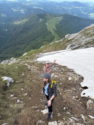 Two women are standing on a rocky, grassy slope in the mountains near Jošanica, Montenegro. The woman in the foreground is wearing a black and white plaid shirt, a blue t-shirt, black pants, and a leopard-print baseball cap. She has a yellow backpack on. The woman behind her is wearing a pink hooded sweatshirt, a pink baseball cap, sunglasses, and is holding a cell phone. She is raising her right hand. A patch of snow is visible to their right. In the background, the mountains are covered in dense green forest, with a winding dirt road visible in the distance. The sky is overcast.