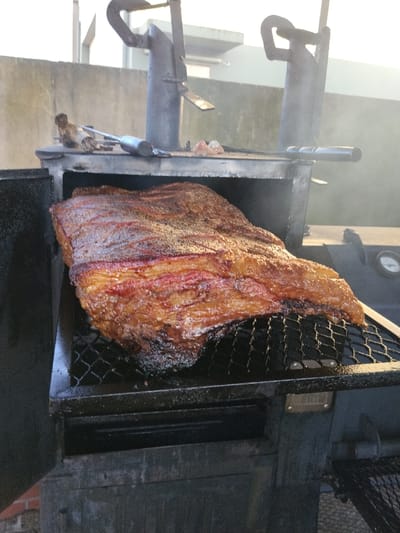 A large piece of cooked meat sits on a black grill inside a smoker. The meat has a dark, crispy exterior with visible layers of pink within. The smoker is made of dark metal with two vertical chimney stacks and a temperature gauge on the side. A concrete wall and smoke are visible in the background.