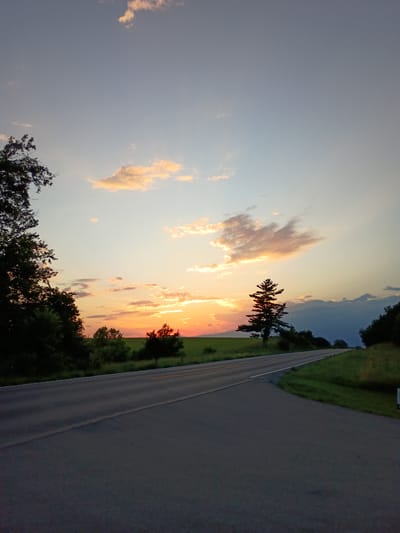 A rural road curves through a grassy landscape under a twilight sky. Silhouetted trees line the left side of the road, with a few scattered clouds tinged with orange from the setting sun. In the distance, a solitary evergreen tree stands against the horizon. The road surface is asphalt, with a white line marking the center. The sky transitions from a pale blue at the top to warmer hues near the horizon. No people or vehicles are visible in the image.