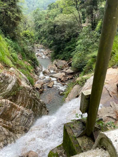 From an elevated perspective near Ramboda, Sri Lanka, a waterfall plunges into a rocky creek, viewed through the railing of a bridge or observation point. Lush, green vegetation covers the rocks, while a densely forested area is visible in the background. The water cascades over the cliff's edge, creating a strong contrast with the darker, layered rock formations. A metal pipe, likely part of the bridge structure, crosses the upper portion of the frame.