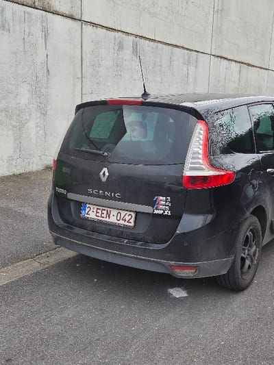 A black Renault Scenic, bearing Belgian license plate 2-EEN-042, is parked on an asphalt surface alongside a textured concrete wall composed of horizontal panels. The vehicle is viewed from its rear-left quarter. A "TURBO" badge is affixed to the lower left of the tailgate, and "SCENIC" is centered above the license plate. A Renault diamond logo is positioned below the rear window. On the lower right of the tailgate, a multi-tiered decal resembling BMW M-series logos displays "M3", "M4", "M5", and "JMP.X1". A rear windshield wiper and a roof-mounted antenna are visible. Through the rear window, a light-skinned individual's face, appearing to be a child or small adult, is discernible, facing towards the rear of the vehicle. The car's black rear wheel is equipped with an alloy rim. The concrete wall behind the car exhibits staining and wear. A curb line is visible on the left side of the asphalt. This scene is situated in the city of Maldegem, Belgium.