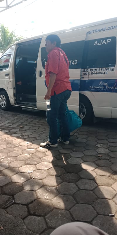 A male individual stands on a hexagonal paver surface in Bojongherang, Indonesia. He wears a red patterned short-sleeved shirt, dark blue jeans, and white athletic shoes. He holds a clear plastic water bottle in his right hand, and a teal duffel bag rests at his left foot. To the right of the man is a white passenger van with its sliding side door open, revealing a dark interior. The van displays "SILIWANGI TRANS SHUTTLE" branding, a tiger logo, the website "NGITRANS.COM", and a phone number "(022) 84468648". Bright sunlight illuminates the scene, casting prominent shadows on the ground. A partial roof structure and foliage are visible in the background.