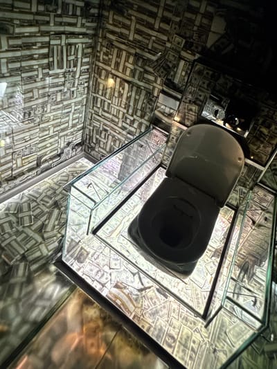 This media file depicts a unique and elaborate bathroom interior, likely a novelty or art installation. The walls, ceiling, and possibly the floor are entirely covered in what appears to be a collage of US dollar bills. The bills are arranged in a visually complex, mosaic-like pattern, creating an overwhelming sense of opulence and perhaps a commentary on wealth.
The central object is a white toilet with its lid open, positioned within a transparent glass enclosure that reflects the dollar-bill decor. The glass box itself adds another layer of visual complexity, multiplying the already abundant imagery of money. The lighting in the scene is artificial, creating a somewhat dim and dramatic atmosphere. A single, soft light fixture is visible on the ceiling, which is also covered in dollar bills.
The overall aesthetic suggests an indoor, possibly private or commercial space, designed for novelty and visual impact. There is no indication of people or any specific activity taking place, beyond the implied use of a bathroom. The location context is given as Denver, United States, which might hint at the specific artistic or cultural influences behind such a design. There is no visible text in the image, other than the imprinted text on the dollar bills themselves. The overall impression is one of extravagant and unusual decoration.