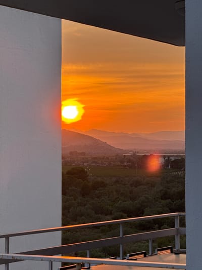 The photograph shows a sunset viewed from what appears to be a rooftop or balcony in Vinaròs, Spain. The sun is positioned low on the horizon, emitting a bright, orange glow. Silhouetted mountains are visible in the background, and a green, forested area extends to the right of the frame. A white wall and a metal structure, possibly part of a railing or ladder, are also in the foreground, framing the sunset scene.