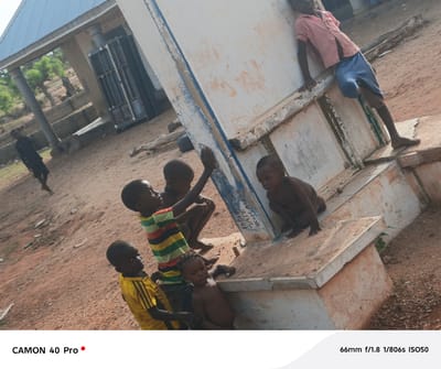 In Gidan Kogi, Nigeria, a group of children are gathered around a concrete structure outdoors. One child in a red shirt and blue shorts leans against the side of the structure. Four other children are closer to the viewer; one wears a yellow and black striped shirt, another a multi-colored striped shirt, and two are shirtless. In the background, an adult in dark clothing walks away from the camera, and a building with a corrugated metal roof and barred windows is visible. The ground is reddish-brown, with scattered debris.