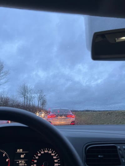 A view from inside a car driving on a rainy day. The windshield is covered in raindrops, blurring the view of the vehicle in front with the license plate "HN:PK 178". Bare trees line the side of the road under a cloudy sky. The car's dashboard is visible, showing a speedometer with a needle pointing around 80 and a digital clock displaying 8:39. Part of the rearview mirror is also visible.