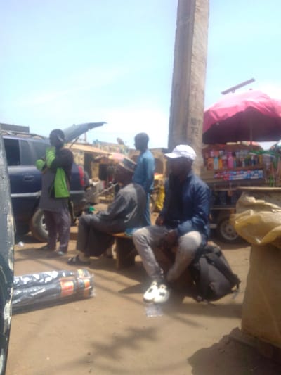 This image captures an outdoor street scene in Mista Ali, Nigeria, under a clear, bright blue sky. The ground consists of bare, light brown dirt.
On the far left, a dark-colored SUV is partially visible, with its rear hatch open. A male subject stands near the SUV, wearing dark clothing and a bright lime-green garment draped over his left shoulder. In the immediate foreground, several black cylindrical bundles, wrapped in clear plastic, rest on the dirt.
Three other male subjects are positioned centrally and to the right. One male, wearing dark traditional attire and sandals, is seated on a plain wooden bench. Behind him, a second male stands, dressed in a light blue long-sleeved shirt and dark trousers. To their right, a third male sits on a dark backpack placed on the ground; he wears a dark blue long-sleeved shirt, light grey trousers, a white baseball cap, and white sneakers with black accents.
On the right side of the frame, a market stall is visible, featuring a red umbrella for shade. The stall displays numerous small bottles and sachets, likely personal care or household products. A tall, slender concrete pillar, marked with "NIPP 32", stands prominently in the background behind the market stall. A large, light-colored sack is partially visible on the extreme right in the foreground.